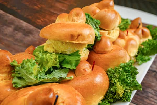 Platter of chicken salad sandwiches on braided rolls with leafy lettuce, garnished with fresh kale.