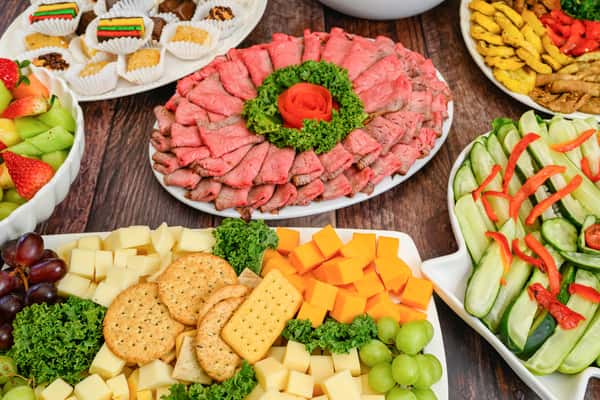 Assorted party platter with roast beef, sliced cheeses, crackers, fresh fruit, desserts, and cucumber salad with red pepper strips.
