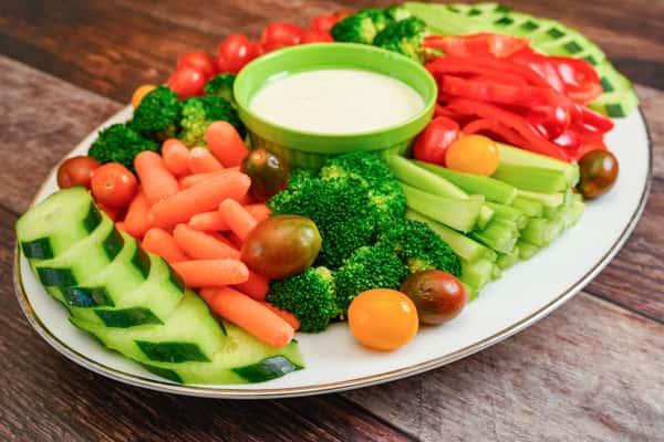 Colorful veggie platter with broccoli, celery, cherry tomatoes, cucumber, carrots, red bell pepper, and a ranch dip in the center.