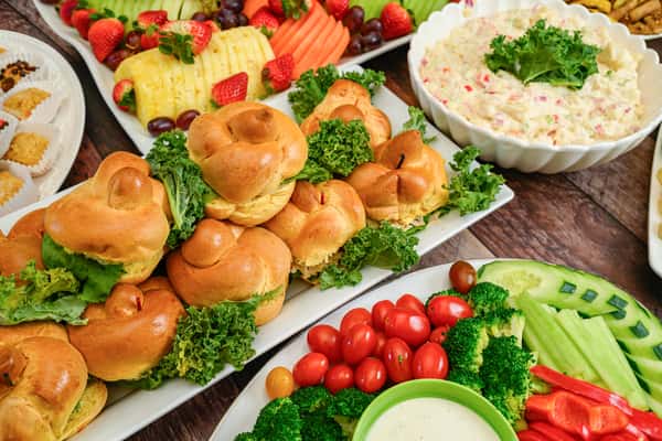 Party platter with mini sandwiches, fresh vegetable tray with dip, fruit slices, and a bowl of creamy potato salad on a wooden table.