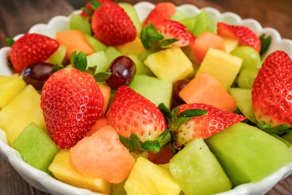 Fresh fruit salad in a white scalloped bowl featuring chunks of honeydew, cantaloupe, pineapple, green grapes, red grapes, and whole strawberries.