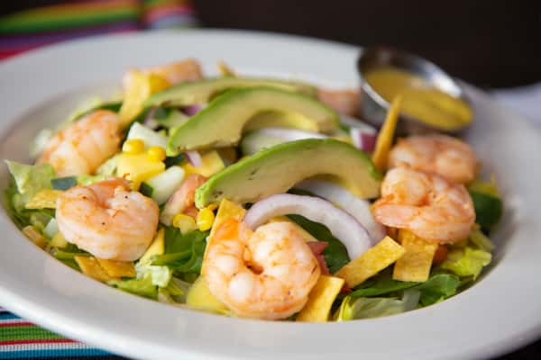 A fresh Vallarta salad featuring grilled shrimp, avocado slices, red onion, corn, and crisp greens, served with a side of dressing.