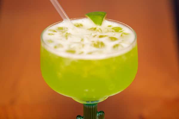 A vibrant green melon margarita served over ice in a large goblet, garnished with a lime wedge and a straw.