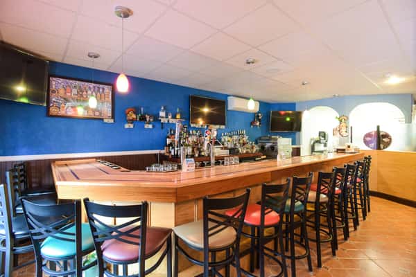 A long, wooden bar counter with black and red stools set against a bright blue wall with multiple television screens and liquor shelving.