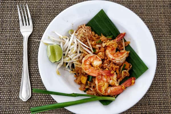 Pad Thai with shrimp, bean sprouts, crushed peanuts, lime wedge, and scallions served on a white plate with banana leaf.