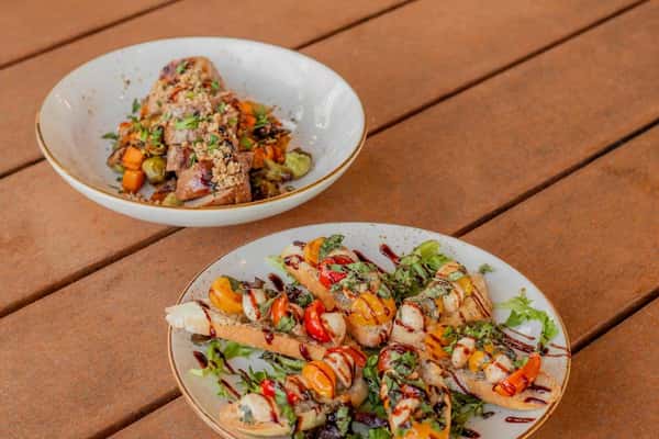 Colorful bruschetta topped with cherry tomatoes, mozzarella, basil, and balsamic glaze next to roasted pork with vegetables.