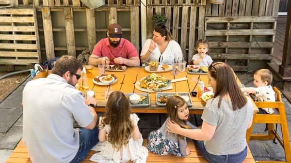Large family enjoying an outdoor meal together around a wooden picnic table with various plates of food and drinks.