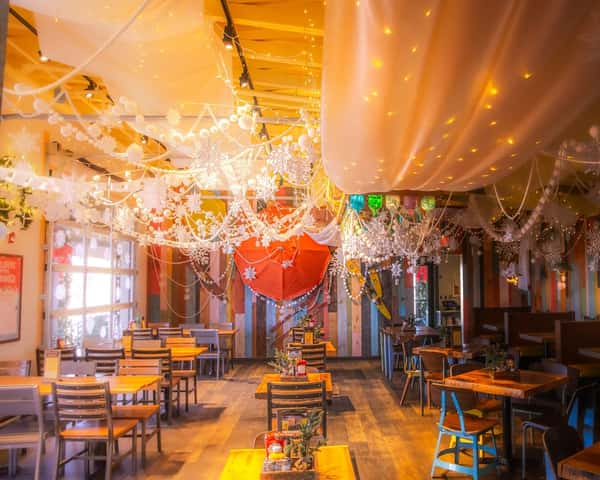 Indoor dining area with wooden tables, festive holiday decor, hanging snowflakes, string lights, and draped white fabric on ceiling.