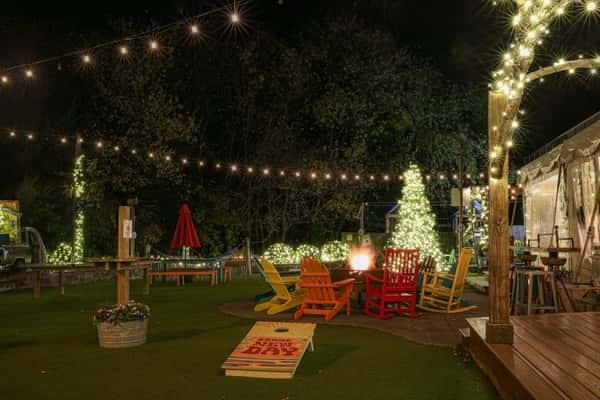 Outdoor fire pit area with colorful chairs, string lights, and glowing holiday trees, featuring a festive cornhole board.