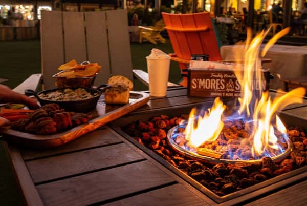 A cozy fire pit table with assorted appetizers, hot drink, and a S'mores Station kit at an outdoor evening dining area.