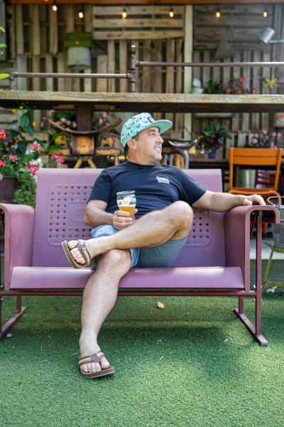 Man in a cap and sandals relaxes on a pink bench with a drink in hand, surrounded by flowers and outdoor decor.