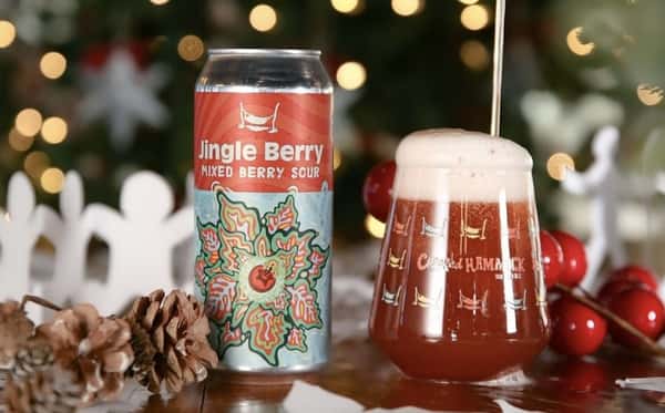 A can and glass of Crooked Hammock's Jingle Berry mixed berry sour beer with pinecones and holiday lights in the background.