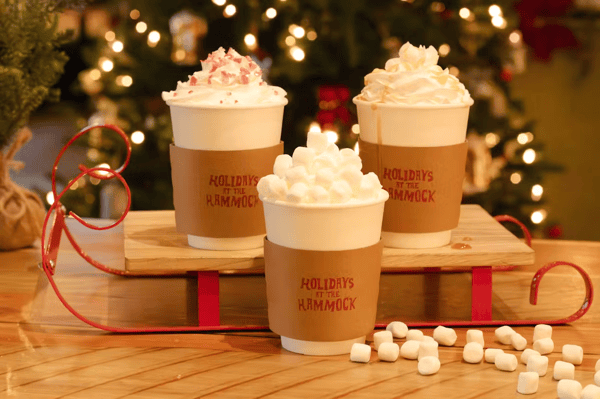 Three festive hot‑chocolate cups topped with whipped cream and marshmallows displayed on a miniature red sled.