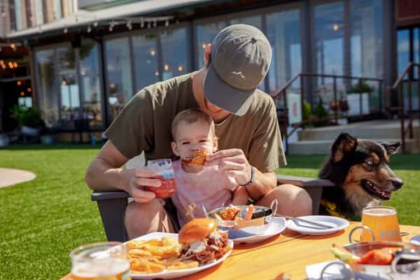 Father feeding baby a chicken wing while sitting outdoors at a picnic table with food and drinks, dog beside them on the grass.