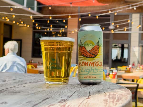 A full pint glass and a can of Hammock Light Lager on a rustic outdoor table at a brewery patio with string lights overhead.