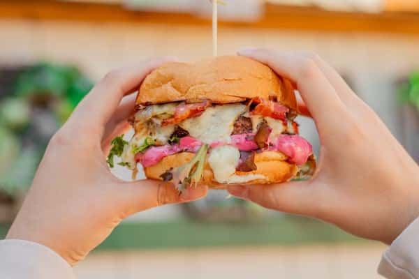 A person holds a loaded burger with melted cheese, bacon, greens, and pink sauce between toasted buns.