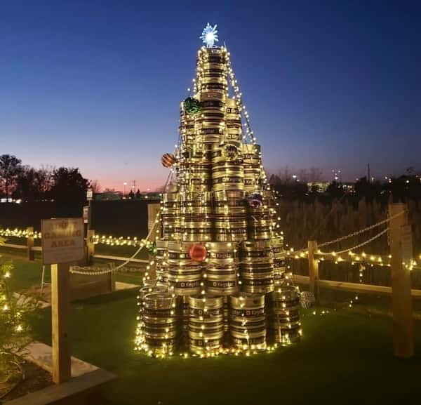 Stacked beer kegs shaped like a Christmas tree, wrapped in string lights and ornaments at dusk.