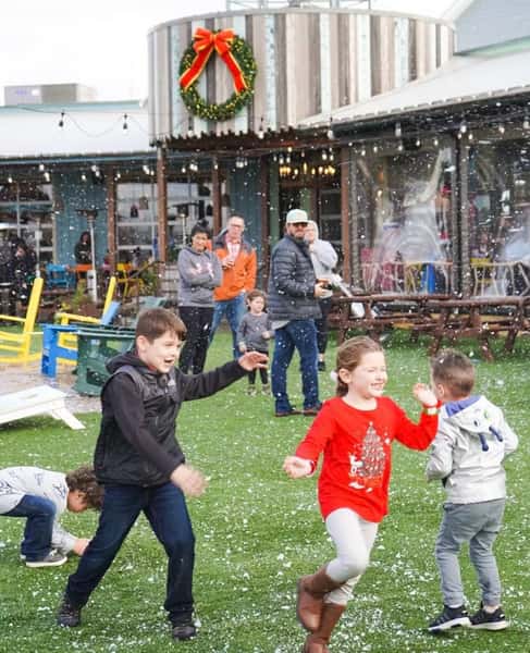 Children playing in artificial snow on a green lawn outside a holiday-decorated building with a large wreath and red bow.