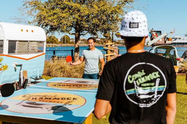 Two men play ping pong on a Crooked Hammock Brewery-branded table near a retro camper and waterfront view.