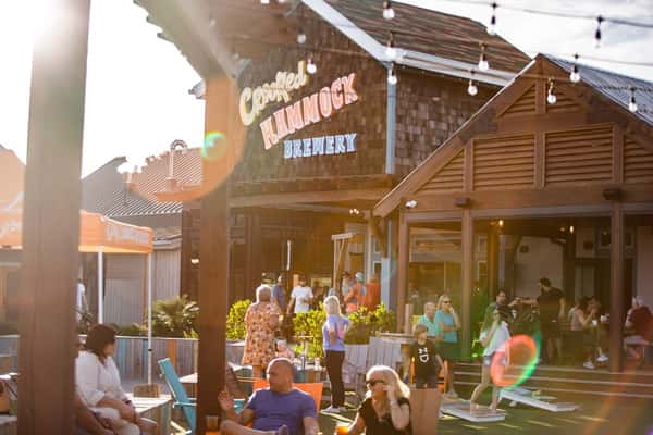 Guests relax and play games outside Crooked Hammock Brewery on a sunny day with lens flare and string lights above.