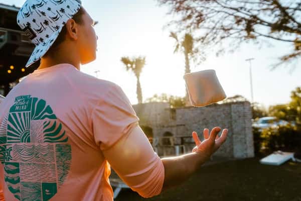 Man in a palm tree hat and pink "Hammock State of Mind" shirt tosses a bean bag outdoors at sunset, ready for a game of cornhole.