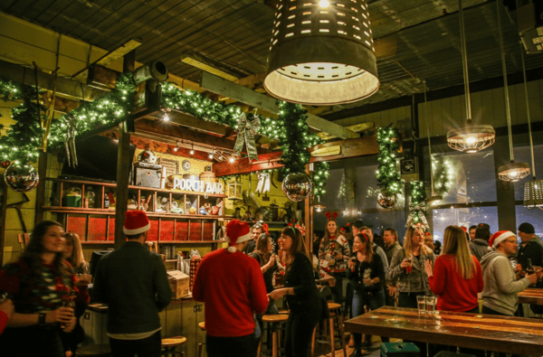 Crowd in festive attire mingling at the Porch Bar decorated with garlands, lights, and ornaments during a holiday celebration.