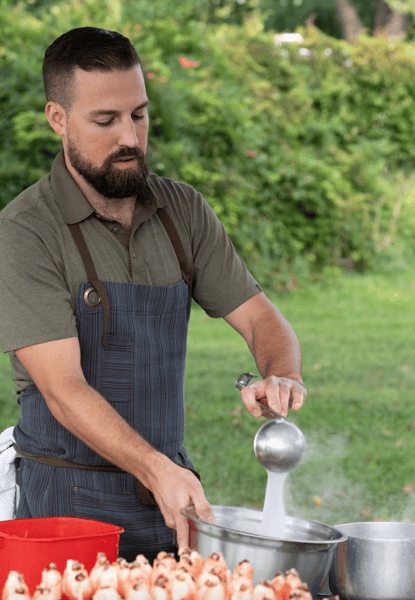 Man in an apron ladles steaming liquid into a bowl at an outdoor food setup with shrimp neatly arranged in front.