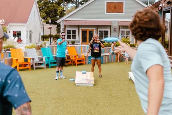 People enjoying a game of cornhole on a lawn surrounded by colorful Adirondack chairs and a cozy outdoor setting.