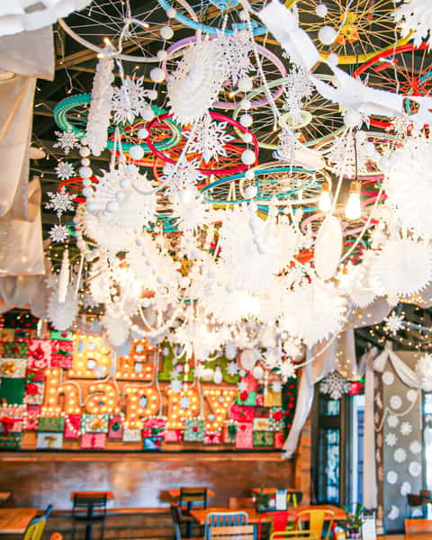 Ceiling decorated with white snowflakes, pom-poms, and bicycle wheels above a festive "Be Happy" holiday-themed dining area.