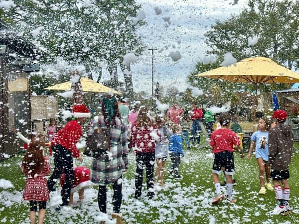 Children and adults play on a grassy lawn under artificial snow with festive outfits and umbrellas creating a cheerful winter scene.