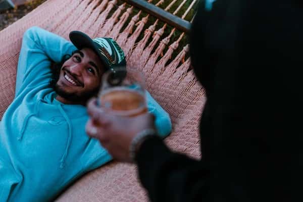Man in blue hoodie smiling while relaxing in a hammock, looking up at a person holding a drink in the foreground.