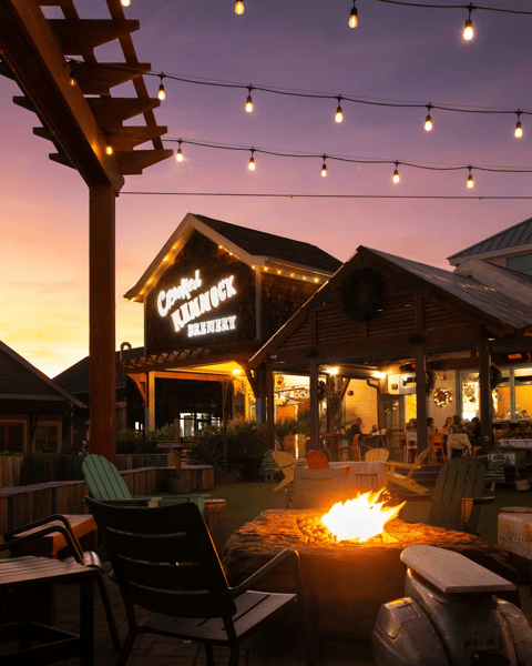 Fire pit surrounded by chairs at Crooked Hammock Brewery with string lights glowing at sunset.