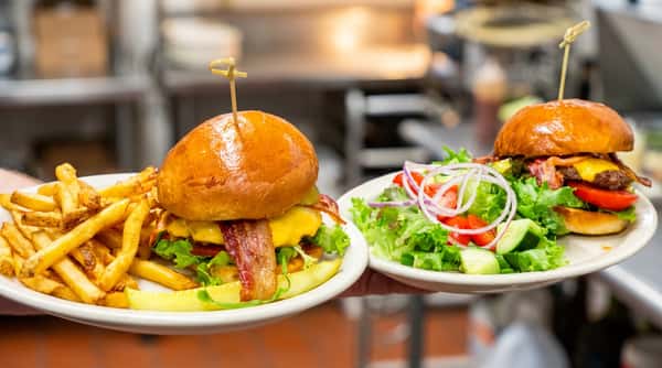 Person in blue "Matty B's" shirt holding two burgers: one with fries and bacon, the other with a side salad.