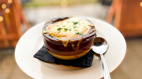 French onion soup in a brown bowl with melted cheese and parsley, on a white plate with a spoon.