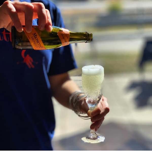 A person in a blue shirt pouring Mionetto Prosecco into a fluted glass, filling it with bubbly, pale yellow liquid.