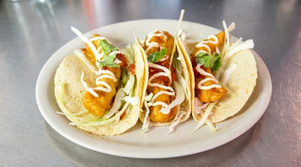 Three fish tacos on corn tortillas with shredded cabbage, salsa, breaded fish, crema, and cilantro, served on a white plate.