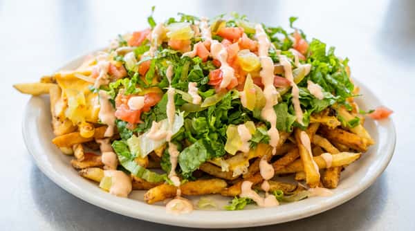 Loaded fries with melted cheese, chopped lettuce, diced tomatoes, pickles, and a drizzle of pink sauce on a white plate.