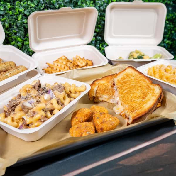Takeout containers with waffle fries, mac and cheese, and a grilled sandwich on a wooden table