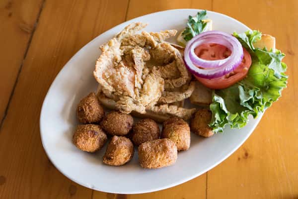 A golden-brown fried fish fillet is served on a white plate alongside crispy hush puppies, a small cup of pink dipping sauce, sliced red onion, and a garnish of fresh parsley. The dish is presented simply, highlighting a classic seafood meal with a crispy, freshly fried finish.