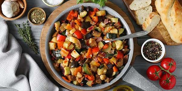 Roasted vegetable ratatouille with zucchini, eggplant, bell peppers, and tomatoes, served in a rustic bowl with sliced baguette and fresh herbs on a wooden table.