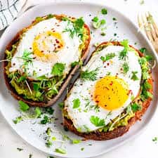 Avocado toast with sunny-side-up eggs and fresh herbs on rustic bread, plated for a healthy brunch