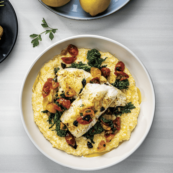 Seared white fish over creamy polenta, topped with sautéed broccolini, blistered cherry tomatoes, and herbs in a modern plated presentation.