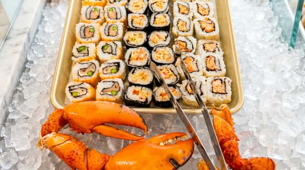 Assortment of sushi rolls with salmon, crab, and avocado next to bright orange cooked lobster claws on ice.