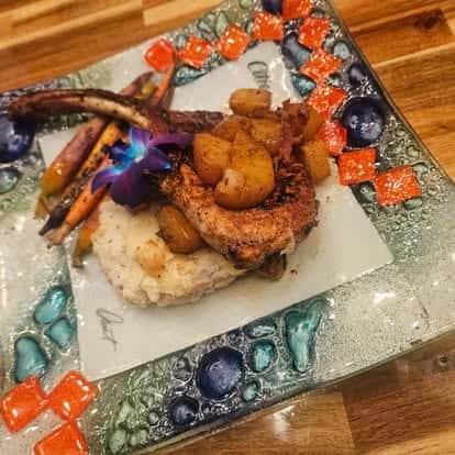 Tomahawk pork chop served over mashed potatoes with roasted root vegetables and spiced apples, garnished with a purple flower on a decorative mosaic plate.