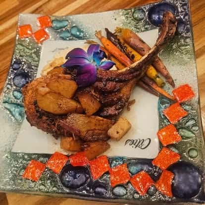 Tomahawk pork chop served with glazed apples, roasted carrots, and root vegetables, garnished with a purple orchid on a colorful mosaic plate.