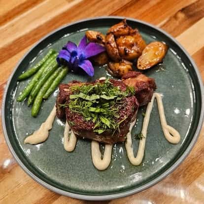 Herb-crusted lamb chops drizzled with aioli, served with roasted fingerling potatoes, green beans, and a vibrant purple orchid garnish on a green ceramic plate.