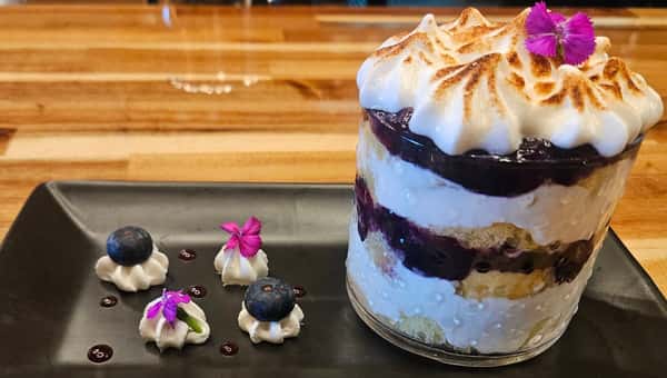 Layered blueberry trifle with whipped cream, cake, and berry compote in a glass cup, garnished with a flower, alongside piped whipped cream and blueberries on a black plate.