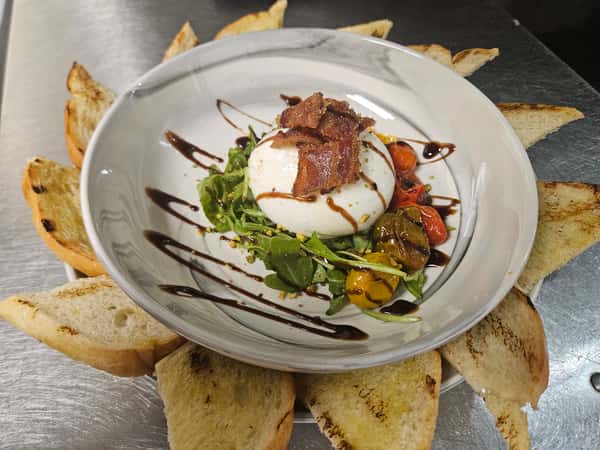 Fresh burrata cheese served over arugula, roasted cherry tomatoes, and pesto, topped with crispy bacon and drizzled with balsamic glaze, surrounded by grilled crostini on a white plate.