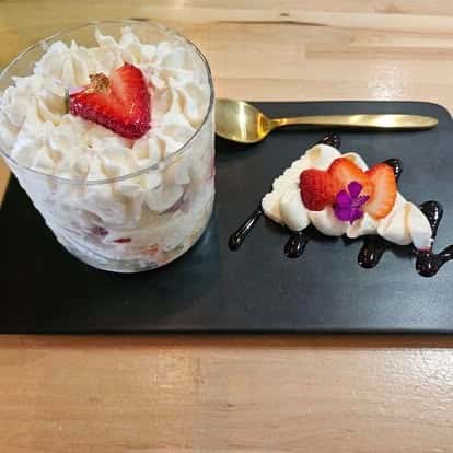 Strawberry shortcake parfait with whipped cream in a glass cup, served alongside a decorative swirl of whipped topping, sliced strawberries, and chocolate sauce on a black plate with a gold spoon.