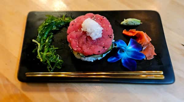 Tuna tartare stack topped with sushi rice, accompanied by seaweed salad, pickled ginger, wasabi, and a blue orchid garnish, plated on a black rectangular dish with gold chopsticks.